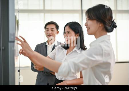 Una giovane operatrice asiatica di marketing, attraente e sicura, ha condiviso le sue idee con i suoi colleghi mentre guardava le note appiccicose su un tabellone Foto Stock