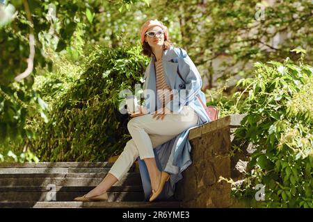 Una donna felice seduta tra le piante, sorridente in abbigliamento casual alla moda Foto Stock