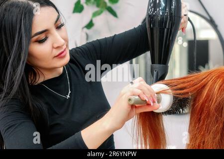 Bella donna stilista fare capelli styling con asciugacapelli al cliente in moderno salone di bellezza. Il parrucchiere rende l'acconciatura per donna caucasica di mezza età con capelli rossi. Foto Stock