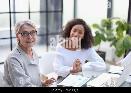 Due donne in viaggio sorridono mentre lavorano insieme su un computer portatile a un tavolo nella sala riunioni dell'ufficio Foto Stock