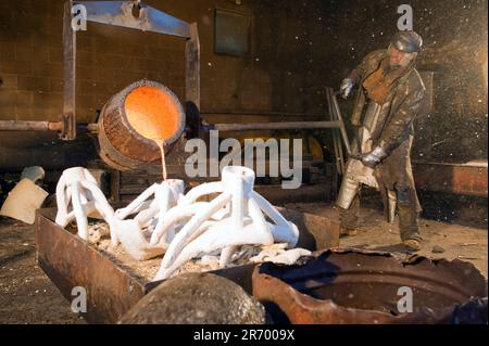 Un uomo che versa bronzo fuso arancione in modanature bianche in una fonderia con l'aiuto di una gru di supporto. Foto Stock