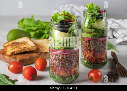 Insalata sana con quinoa, avocado, pomodoro ciliegia, ravanello, spinaci, E olio d'oliva in vasetti di Mason su fondo grigio Foto Stock