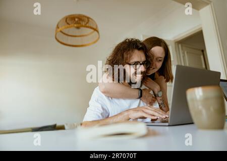 Uomo sorridente che lavora sul computer portatile mentre moglie lo abbraccia a casa. Concetto di lavoro a distanza Foto Stock