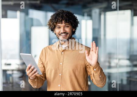 Ritratto di giovane programmatore con computer tablet all'interno dell'ufficio, uomo ispanico con capelli ricci sorridente e guardando la macchina fotografica, uomo sul posto di lavoro che prova applicazioni online in piedi vicino alla finestra. Foto Stock