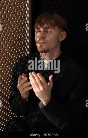 Sacerdote cattolico che prega vicino alla finestra di legno in cabina confessionale Foto Stock