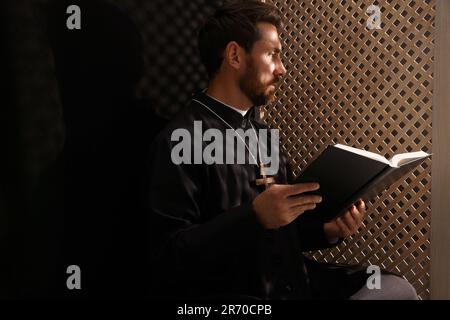 Sacerdote cattolico in cassock leggendo la Bibbia in cabina confessionale Foto Stock