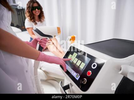 Vista ravvicinata mani di estetista in guanti rosa, che tiene il sensore laser con una mano e che regola il dispositivo laser sul tabellone con l'altra. Client soddisfatto in sfondo sfocato. Foto Stock