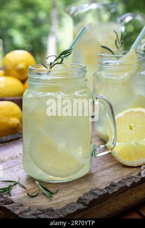 Limonata in due gialle di vetro con limoni a fette, rosmarino e ghiaccio all'aperto su un vassoio di legno Foto Stock