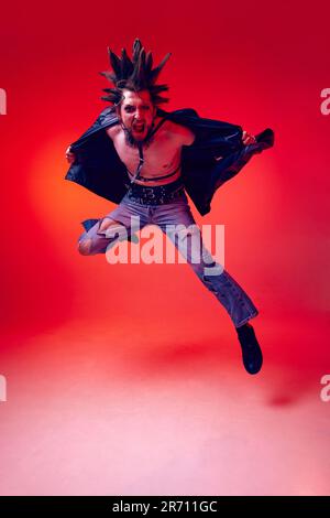 Ritratto a tutta lunghezza di un giovane punk in abiti straordinari, acconciature e trucco che si posano su sfondo rosso studio in luce al neon Foto Stock