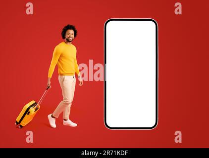 Un viaggiatore con un bagaglio giallo a valigia che cammina vicino a uno smartphone gigantesca con schermo vuoto. Sorridente afro-americano spensierato ragazzo isolato su rosso raccomanda app per viaggi e viaggi Foto Stock