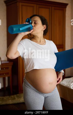 Donna incinta beve acqua, rinnovando l'equilibrio dell'acqua dopo l'allenamento, tenendo il tappetino yoga, godendo di uno stile di vita sano e attivo Foto Stock
