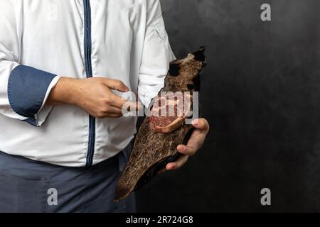 Raccolto anonimo maschio in uniforme che tiene pezzo di carne fresca deliziosa su tavola di legno contro grunge sfondo nero Foto Stock