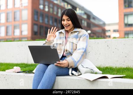 Ragazza entusiasta dello studente che agita la mano al computer portatile mentre ha lezioni online, videochiamate con l'insegnante, seduto nel parco Foto Stock