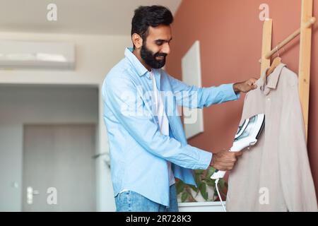 Happy Arabic Man usando il sistema di cottura a vapore dei vestiti in piedi all'interno domestico Foto Stock