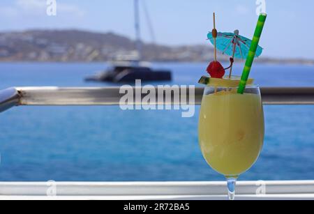 Un grande bicchiere rinfrescante di Pina Colada, decorato con una paglia e un ombrellone con un bel mare e uno yacht di lusso da sfondo sfocato Foto Stock