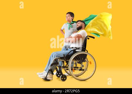 Il ragazzino e il padre in sedia a rotelle indossano cappe supereroi su sfondo giallo Foto Stock