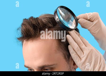 Medico con lente d'ingrandimento che esamina i capelli del giovane uomo su fondo blu, primo piano Foto Stock