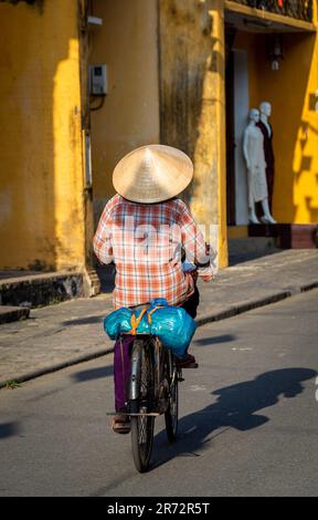La vista posteriore di una donna vietnamita coperta da un cappello di riso di bambù in bicicletta passa le vecchie case con parete gialla in una giornata di sole. Foto Stock