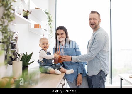 Ritratto di due bicchieri da tostare per adulti e bambini con succo di frutta e sorridente alla macchina fotografica. Simpatica famiglia caucasica gustando frullati freschi fatti in casa durante una dieta sana a casa. Foto Stock