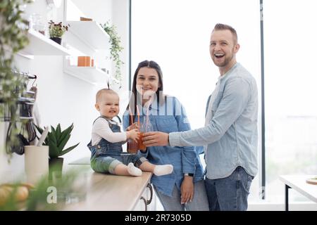 Ritratto di due bicchieri da tostare per adulti e bambini con succo di frutta e sorridente alla macchina fotografica. Simpatica famiglia caucasica gustando frullati freschi fatti in casa durante una dieta sana a casa. Foto Stock