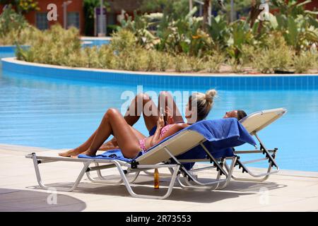 Coppia abbronzata sdraiata su sedie a sdraio e solarium vicino alla piscina. Donna che usa lo smartphone, vacanza in famiglia Foto Stock