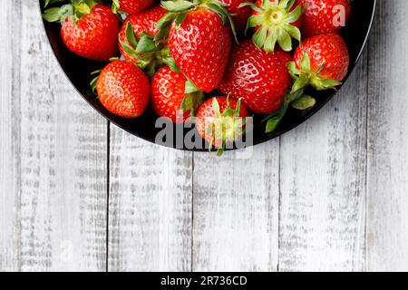 Fragole fresche biologiche in un piatto rotondo nero su uno sfondo di legno chiaro, vista dall'alto, con spazio per il testo. Foto Stock