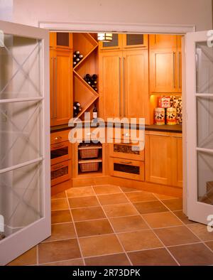 Cabina armadio personalizzata vista attraverso porte a vetri, con mobili in legno caldo, deposito vini, pavimento piastrellato e ripostiglio organizzato in cucina. Foto Stock