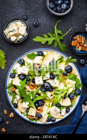 Insalata gourmet con pere dolci, mirtilli, formaggio azzurro, rucola e noci. Sfondo nero del tavolo, vista dall'alto Foto Stock