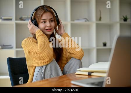 Una giovane donna asiatica attraente in un maglione giallo accogliente ascolta musica o podcast attraverso le sue cuffie mentre si siede al suo tavolo da lavoro a casa. Foto Stock