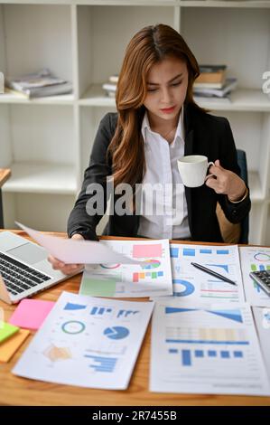 Una donna d'affari asiatica seria e focalizzata giovane o manager finanziario femminile che rivede e analizza i rapporti finanziari aziendali mentre sorseggiano il caffè Foto Stock