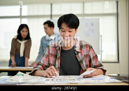 Giovane e felice designer grafico asiatico maschio o UI, lo sviluppatore UX sta rivedendo un nuovo prototipo e lavorando alle sue attività alla sua scrivania. Foto Stock