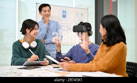 Un giovane e allegro sviluppatore asiatico di sesso maschile seduto al tavolo ha testato un nuovo gioco per la realtà virtuale con occhiali per la realtà virtuale durante la riunione con il suo team. Foto Stock