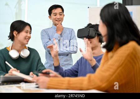 Un allegro sviluppatore asiatico di sesso maschile seduto al tavolo ha testato un nuovo gioco per la realtà virtuale con gli occhiali per la realtà virtuale durante la riunione con il suo team. Realtà virtuale i Foto Stock