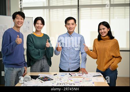 Un gruppo di allegri grafici asiatici o sviluppatori stanno mostrando i loro pollici in piedi durante la riunione insieme. lavoro di squadra, una startup tecnologica Foto Stock