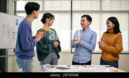 Un gruppo di allegri designer grafici asiatici o sviluppatori è in piedi nella riunione, battendo le mani per celebrare e congratularsi con una coll femminile Foto Stock