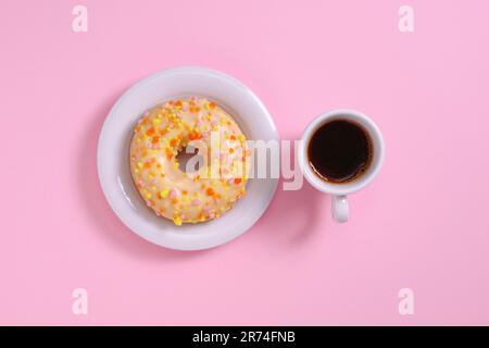 Ciambella con glassa e spruzzata e tazza di caffè nero su sfondo rosa, vista dall'alto Foto Stock