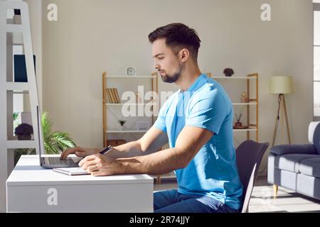 Uomo che lavora a casa o in ufficio su un computer portatile e che scrive alla scrivania prendendo appunti. Foto Stock