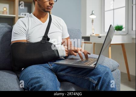 Giovane uomo con un'imbragatura per le braccia sul polso rotto seduto sul divano e al lavoro sul computer portatile Foto Stock