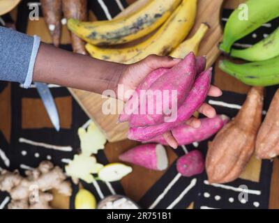 Donna nera che tiene la patata dolce rossa africana, altri ingredienti locali sullo sfondo per la cucina tradizionale ricetta Foto Stock