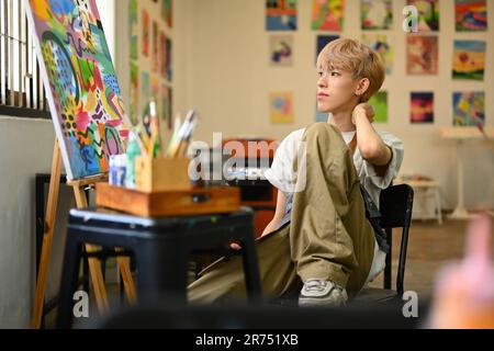 Giovane artista gay pensieroso seduto in studio d'arte e guardando lontano, pensando a nuove idee creative. Arte e concetto di educazione Foto Stock