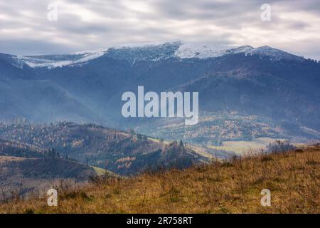 paesaggio di campagna con campi rurali sulle colline. vette innevate delle montagne dei carpazi in lontananza. scenario cupo di cresta borzhava con Foto Stock