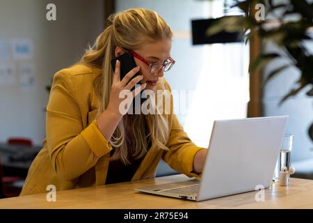 Donna d'affari caucasica casual di taglia più grande con occhiali che usa il computer portatile e parla al telefono alla scrivania Foto Stock