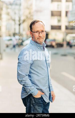 Ritratto all'aperto di bell'uomo di mezza età che indossa una camicia Oxford a manica lunga, che si posa in città, tenendo la mano nella tasca dei jeans Foto Stock