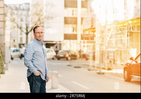 Ritratto all'aperto di bell'uomo di mezza età che indossa una camicia Oxford a manica lunga, che si posa in città, tenendo la mano nella tasca dei jeans Foto Stock