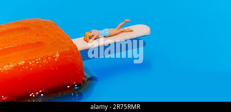 primo piano di una donna in miniatura, con costume da bagno blu, sdraiata sul palo di un melting papsicle arancione su sfondo blu, in forma panoramica Foto Stock