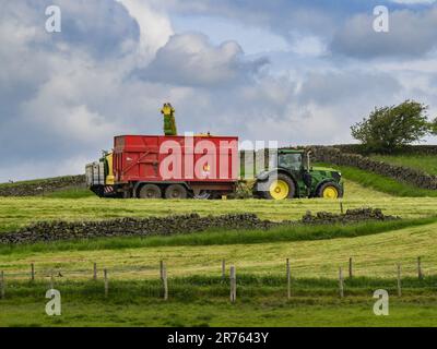 Trattore John Deere 6155R che lavora su terreni agricoli panoramici (riempimento di carri trainati, insilato di erba tagliata, guida di agricoltori) - Inghilterra Regno Unito. Foto Stock