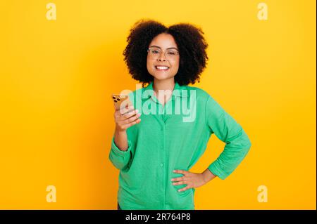 Donna curly afro-americana o brasiliana, vestita camicia verde, tiene il suo smartphone, messaggi con gli amici sui social media, fare ordini online, guarda la fotocamera, sfondo giallo isolato Foto Stock