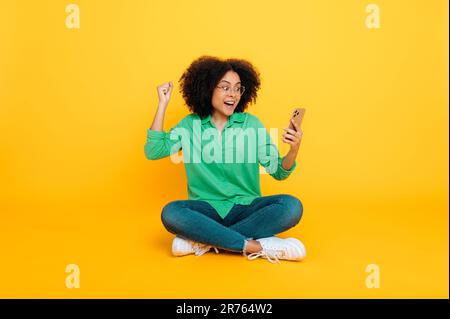 Foto a tutta lunghezza di entusiasta ragazza ricciola afro-americana o brasiliana, siede su uno sfondo giallo, utilizza il suo smartphone, navigando in Internet, si rallegra delle notizie, gestendo le mani, sorrisi Foto Stock