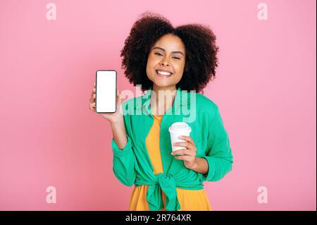 Bella positiva brasiliana o donna ispanica, mostra smartphone con bianco vuoto schermo mock-up per annunci pubblicitari o annunci, contiene una tazza di caffè, sorrisi alla fotocamera, isolato sfondo rosa Foto Stock