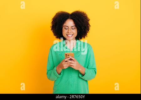 Donna curly afro-americana o brasiliana, vestita maglietta verde, con il suo smartphone, messaggi con gli amici sui social media, fare ordini online, rispondere alle e-mail, sfondo giallo isolato Foto Stock